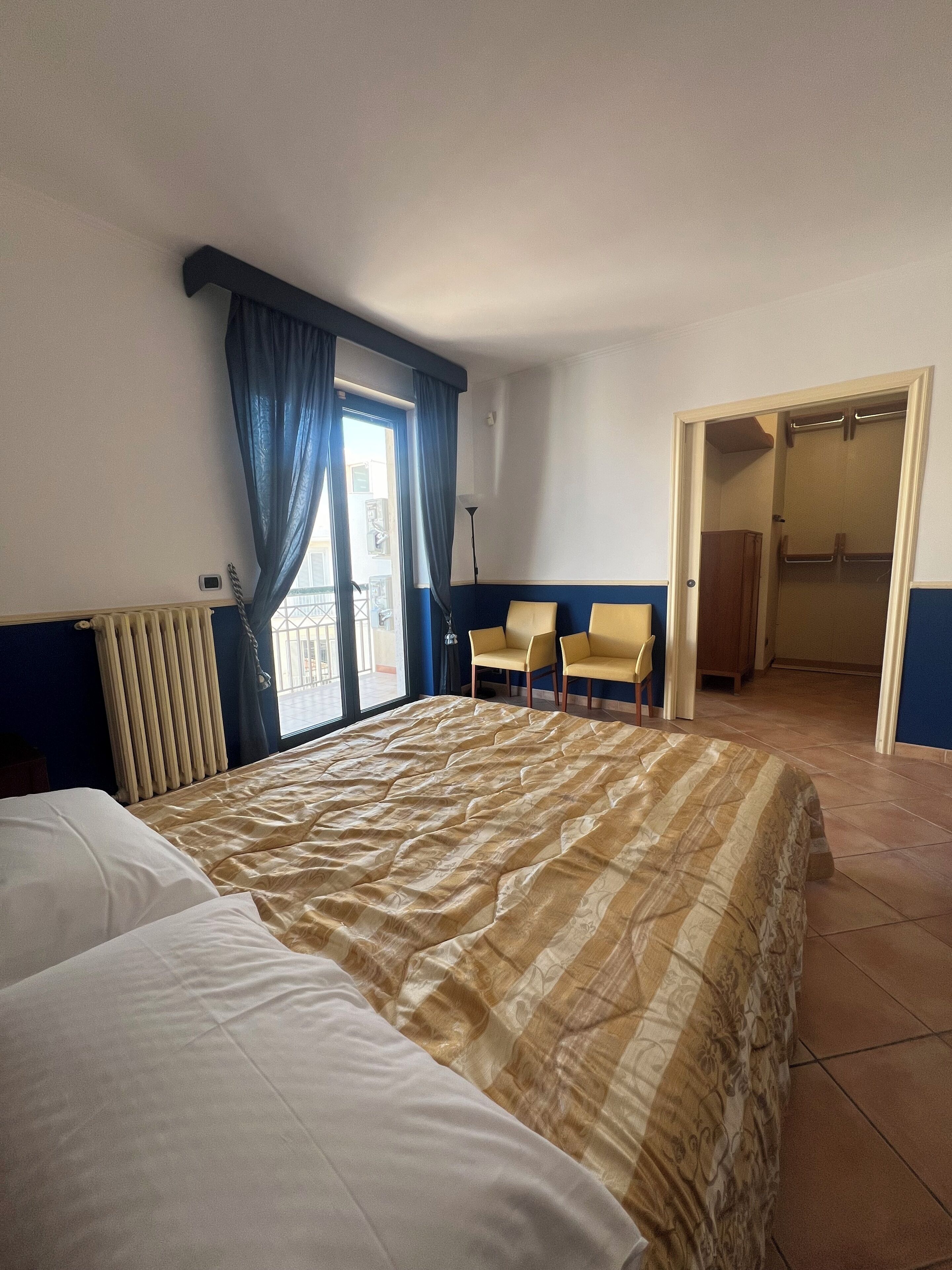 Et hotelværelse med en stor seng, to stole, en radiator, et vindue med gardiner og en åben dør til et skab.