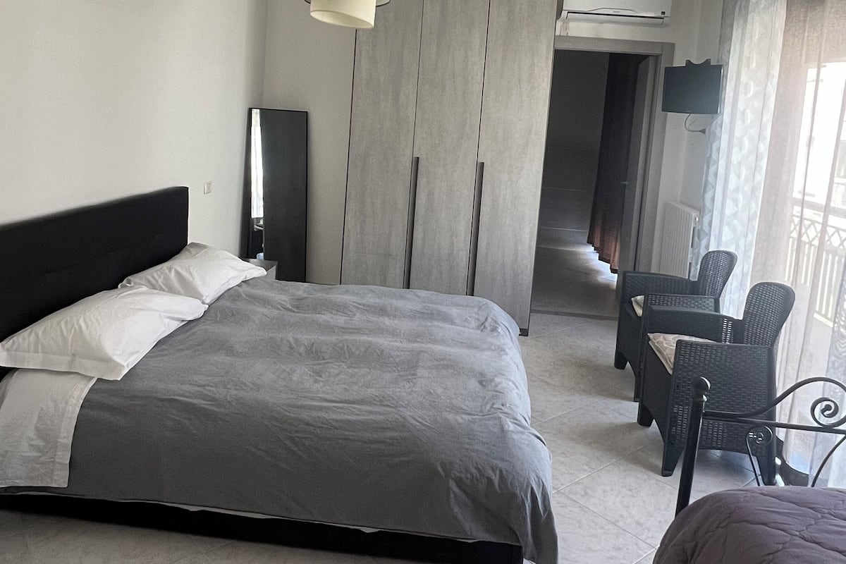 A hotel room with a large bed, a desk with chairs, a TV, and an air conditioning unit.