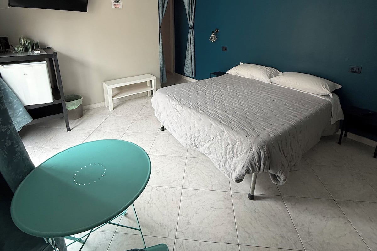 A bedroom with a bed, a desk with a computer, a TV mounted on the wall, and a window with curtains.
