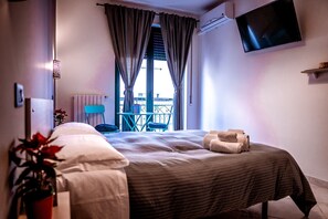 A hotel room with a bed, a TV mounted on the wall, a balcony with a chair, and a potted plant.