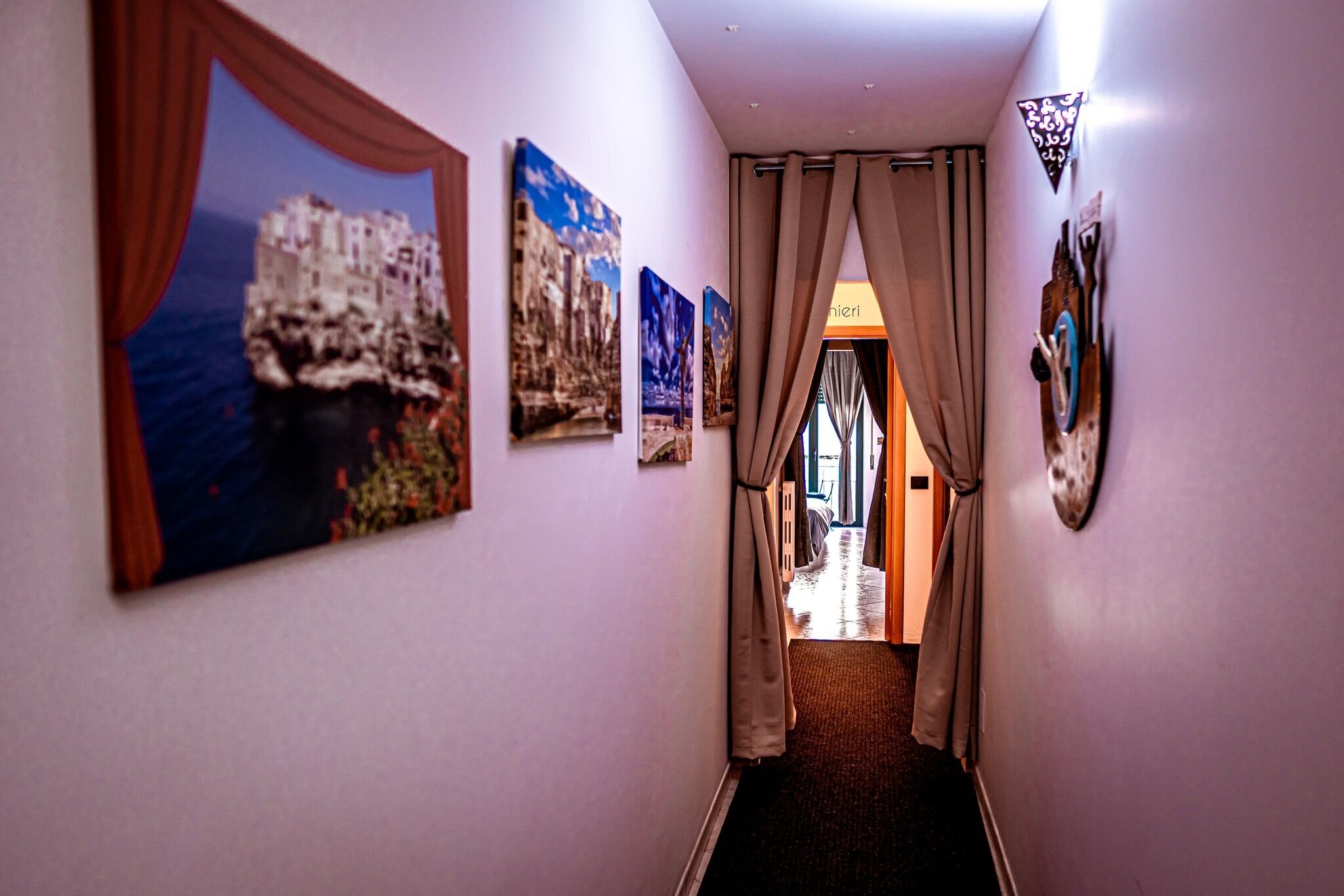 Un couloir qui donne sur une pièce, dans laquelle on peut admirer un grand tableau représentant un paysage côtier, ainsi que plusieurs œuvres d’art plus petites encadrées et un élément décoratif mural.