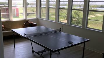 A room with a ping pong table, a ceiling fan, and large windows offering a view of a grassy area and a building.