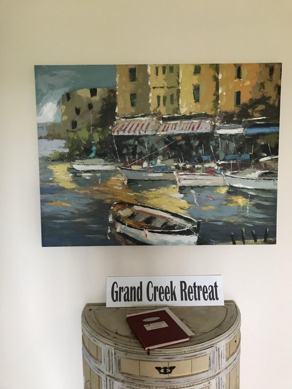 Una pintura de barcos en el agua con edificios al fondo, un letrero que dice 'Grand Creek Retreat' sobre un barril con un libro encima.