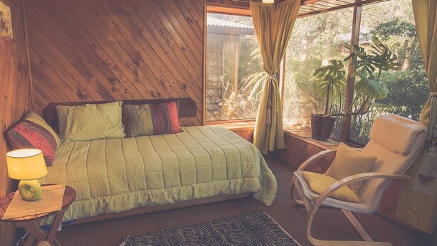 A cozy bedroom with a bed, a rocking chair, a lamp, and a window with curtains. - Silvestre Cabanas (Puerto Varas)