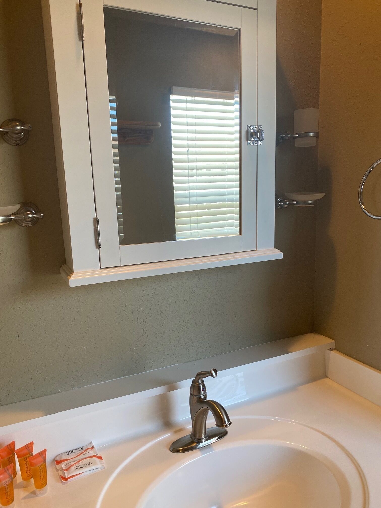Une salle de bain avec un lavabo blanc, un miroir au-dessus et une fenêtre avec des stores.