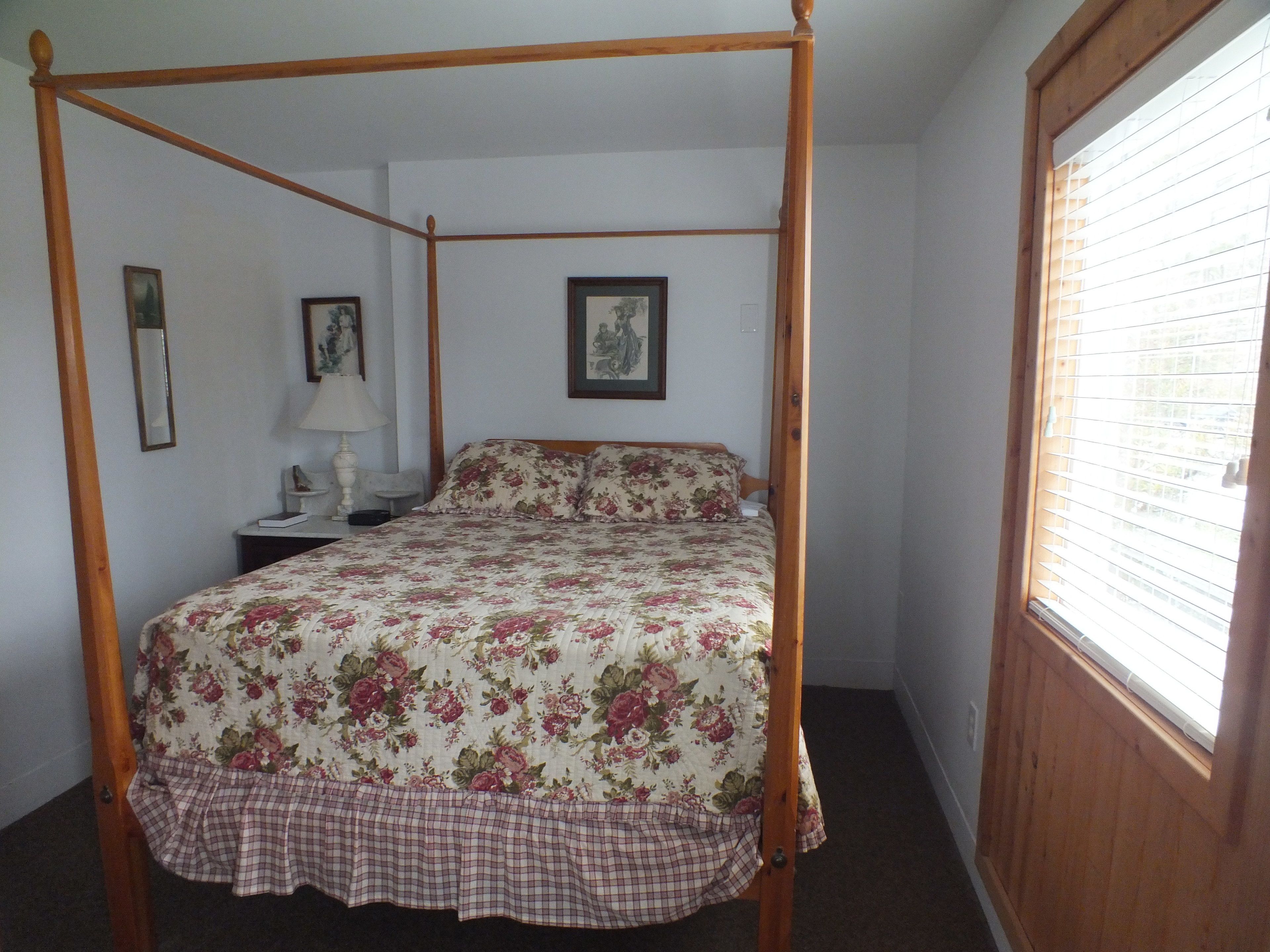 A four-poster bed with floral bedding, a bedside table with a lamp, and framed pictures on the walls.