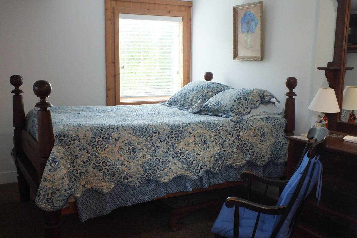 A bedroom with a wooden bed, a window with blinds, a chair, and a bedside table with a lamp.