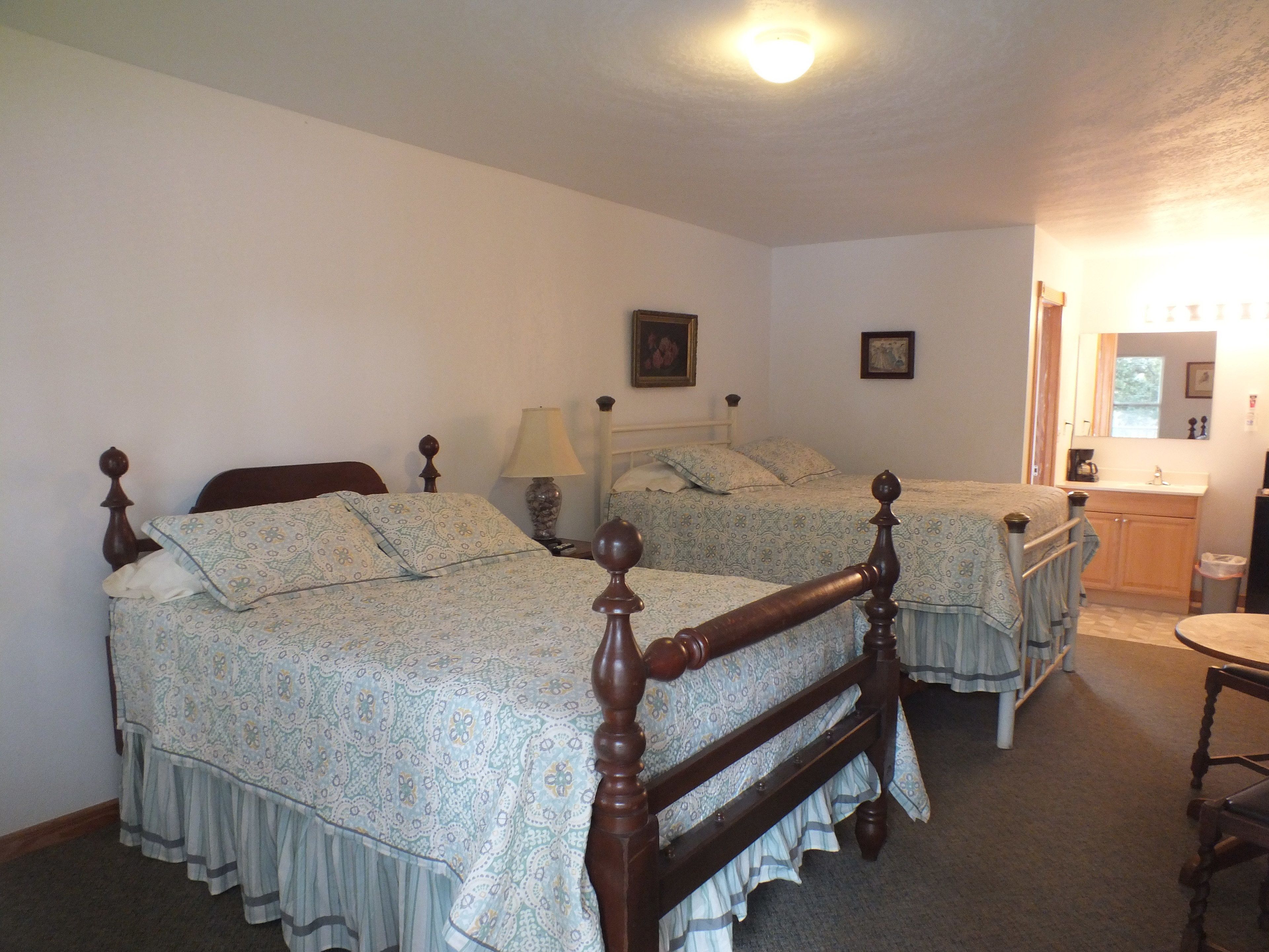 Quarto com duas camas de madeira com colchas franzidas, uma mesa de cabeceira com abajur, cômoda e espelho.