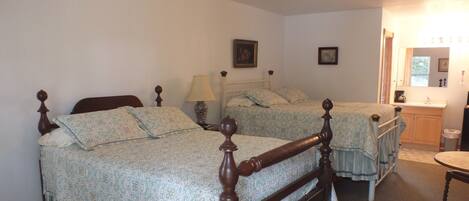 Quarto com duas camas de madeira com colchas franzidas, uma mesa de cabeceira com abajur, cômoda e espelho.