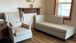 A living room with a wooden floor, a beige armchair, a sofa, a wooden shelf with a vase, and a window with blinds.