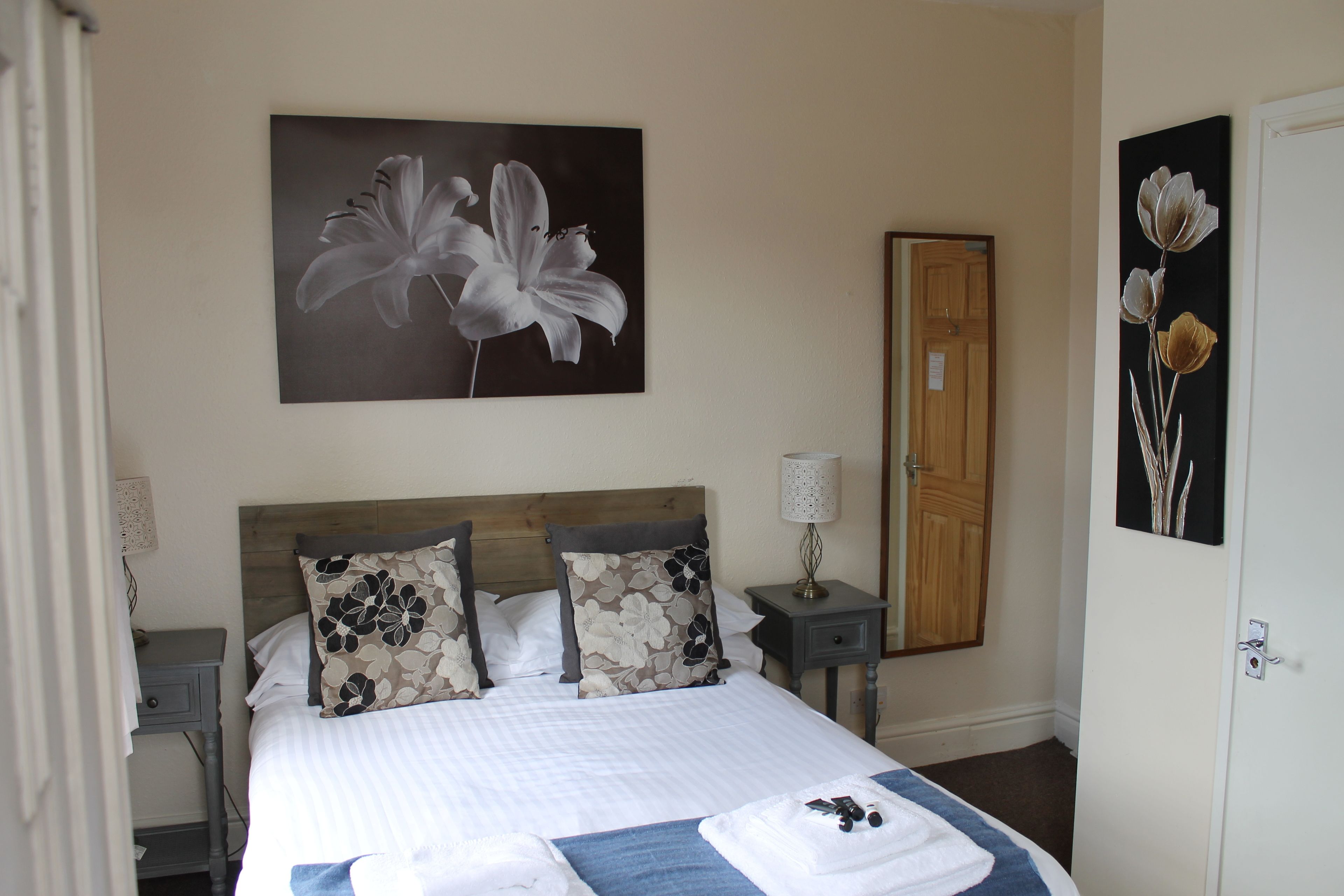 A neatly made bed with floral-patterned pillows, a bedside table with a lamp, and a door to the right.