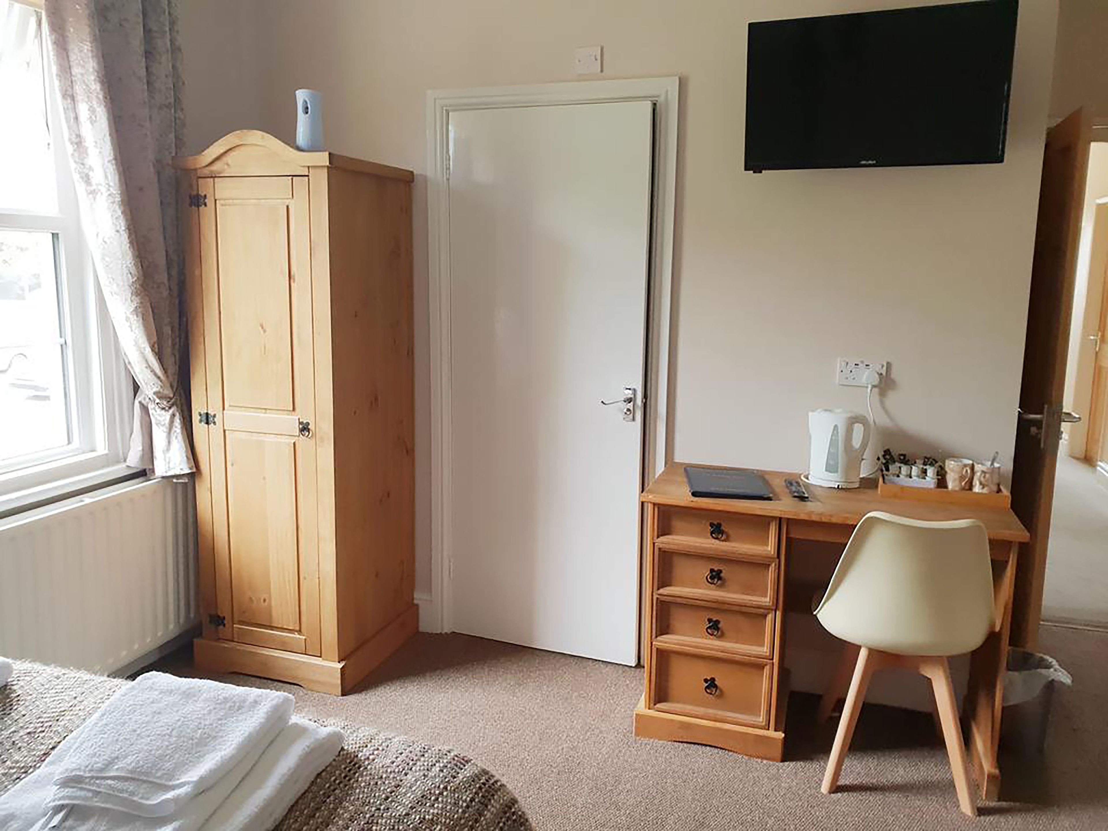 Une chambre avec une armoire en bois, un bureau avec une chaise, un radiateur blanc et un téléviseur fixé au mur.