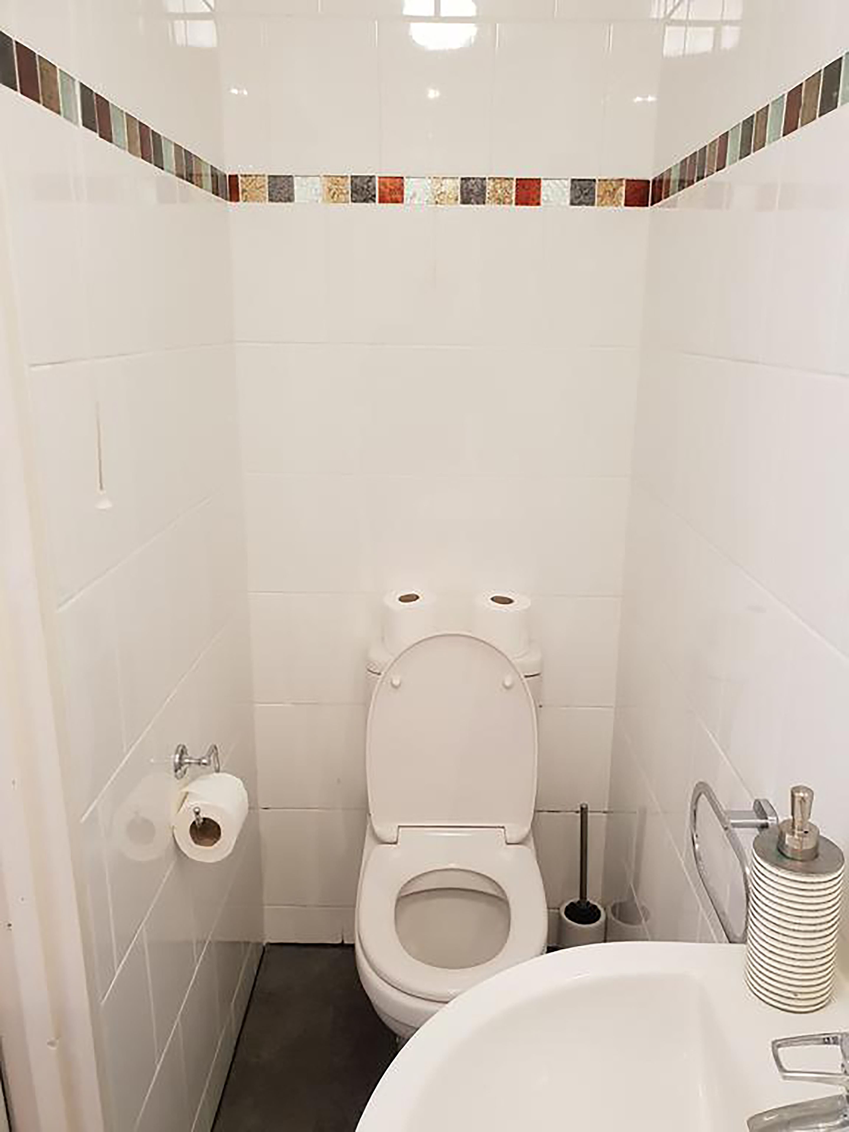 A small, clean bathroom with white tiles, a toilet, a sink, and a toilet paper holder.