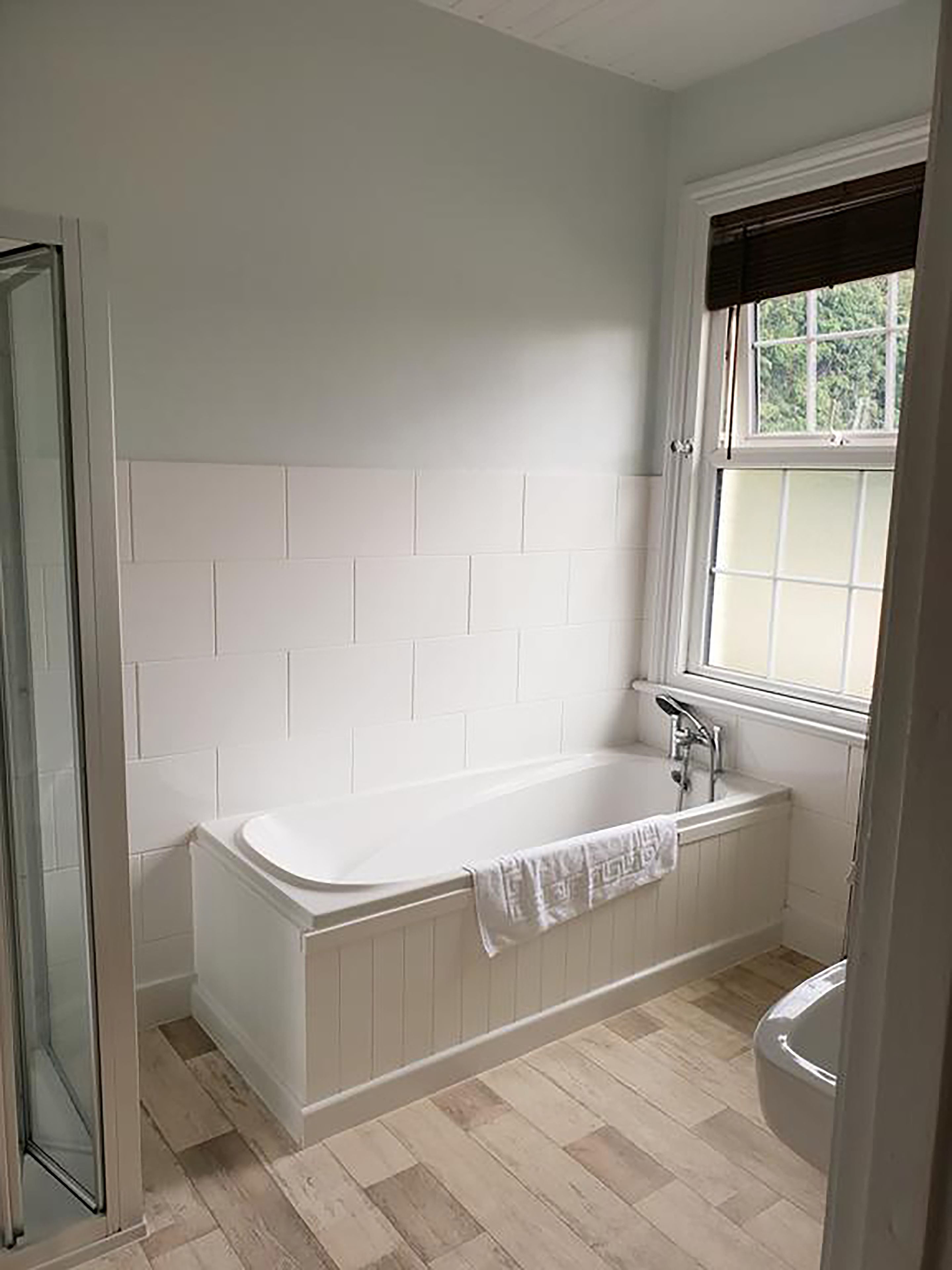 Une salle de bain avec une baignoire blanche, une fenêtre avec des stores et un espace douche.