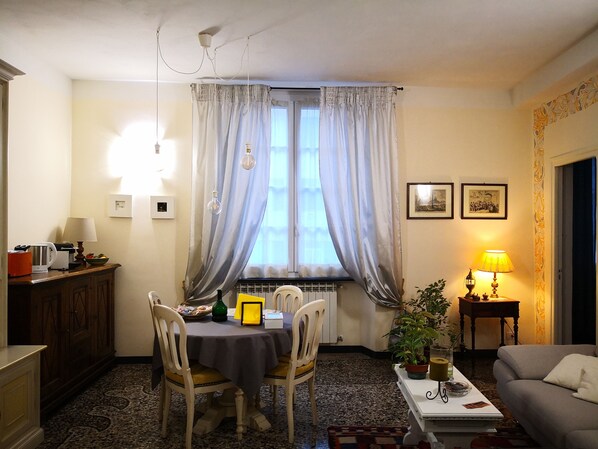 A cozy living room with a dining area, featuring a table set for two, a sofa, a coffee table, and a window with curtains. - B&B Lavagna Uno (Genoa)
