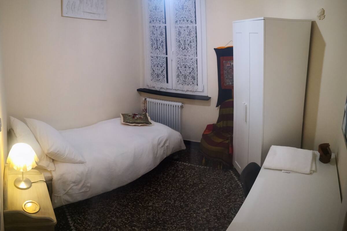 A small, dimly lit bedroom with a single bed, a radiator, a wardrobe, and a small table with a lamp.