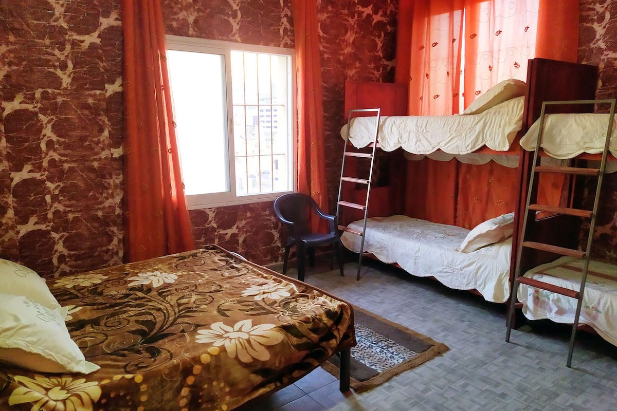 A dormitory room with a bunk bed, a single bed, a chair, and a window with curtains.