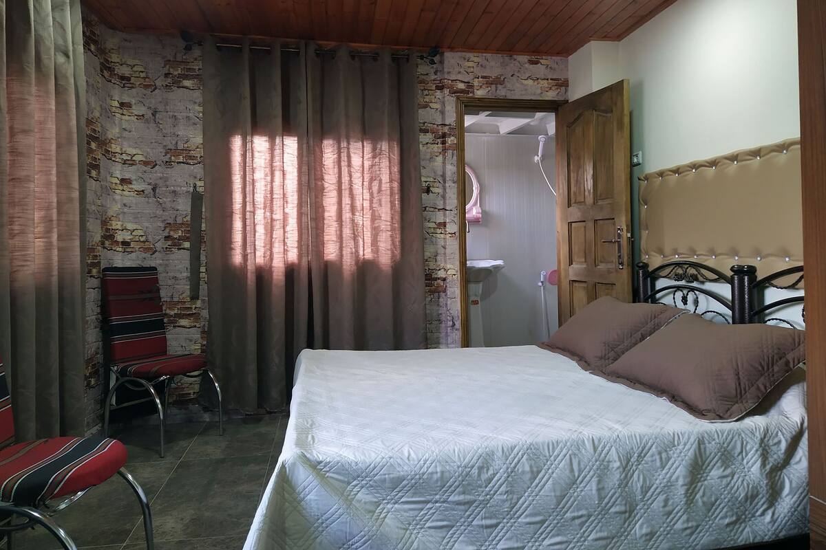 A bedroom with a bed, curtains, a chair, and a wooden ceiling.