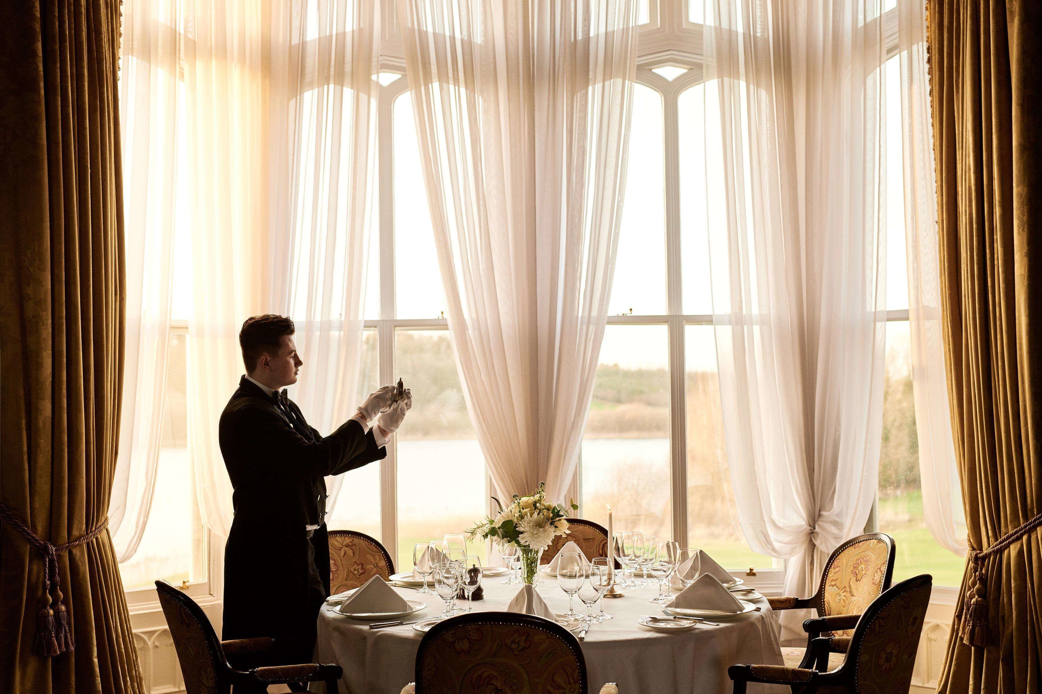 A formal dining room with a table set for a meal, large windows with sheer curtains, and a view of the outdoors.