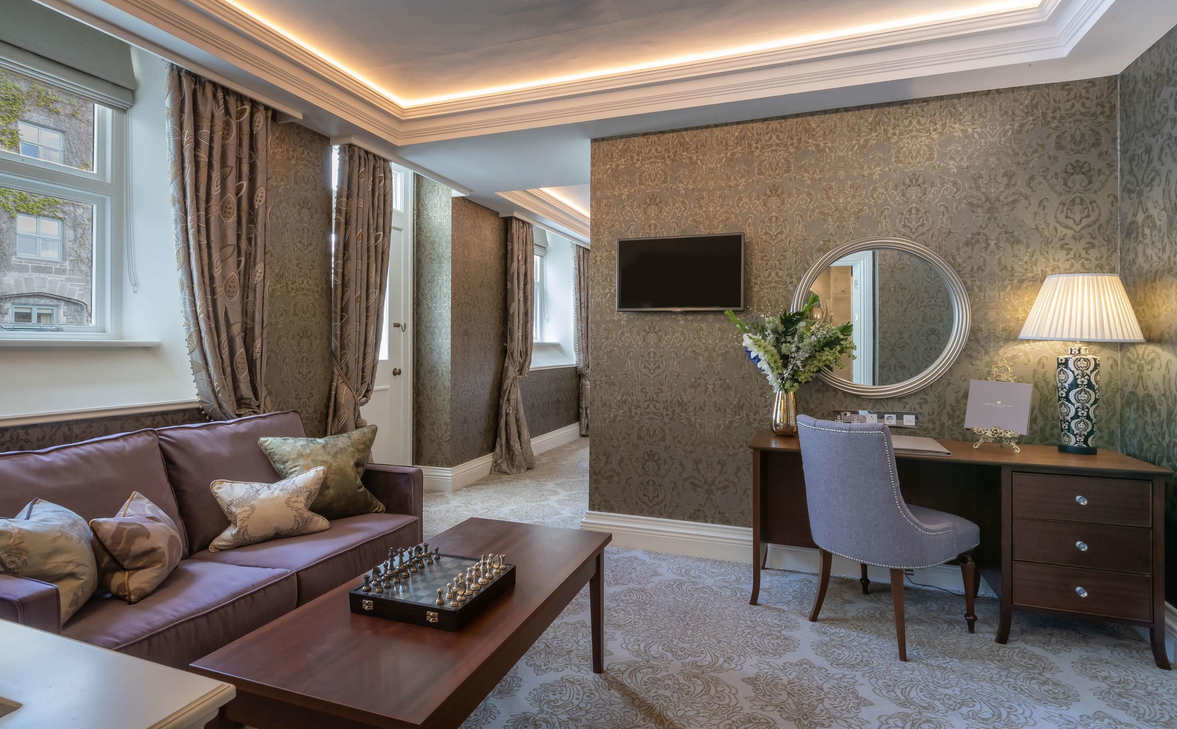 Una habitación con un sofá de cuero marrón, un tablero de ajedrez, un escritorio con una silla, un televisor, un espejo y una lámpara.