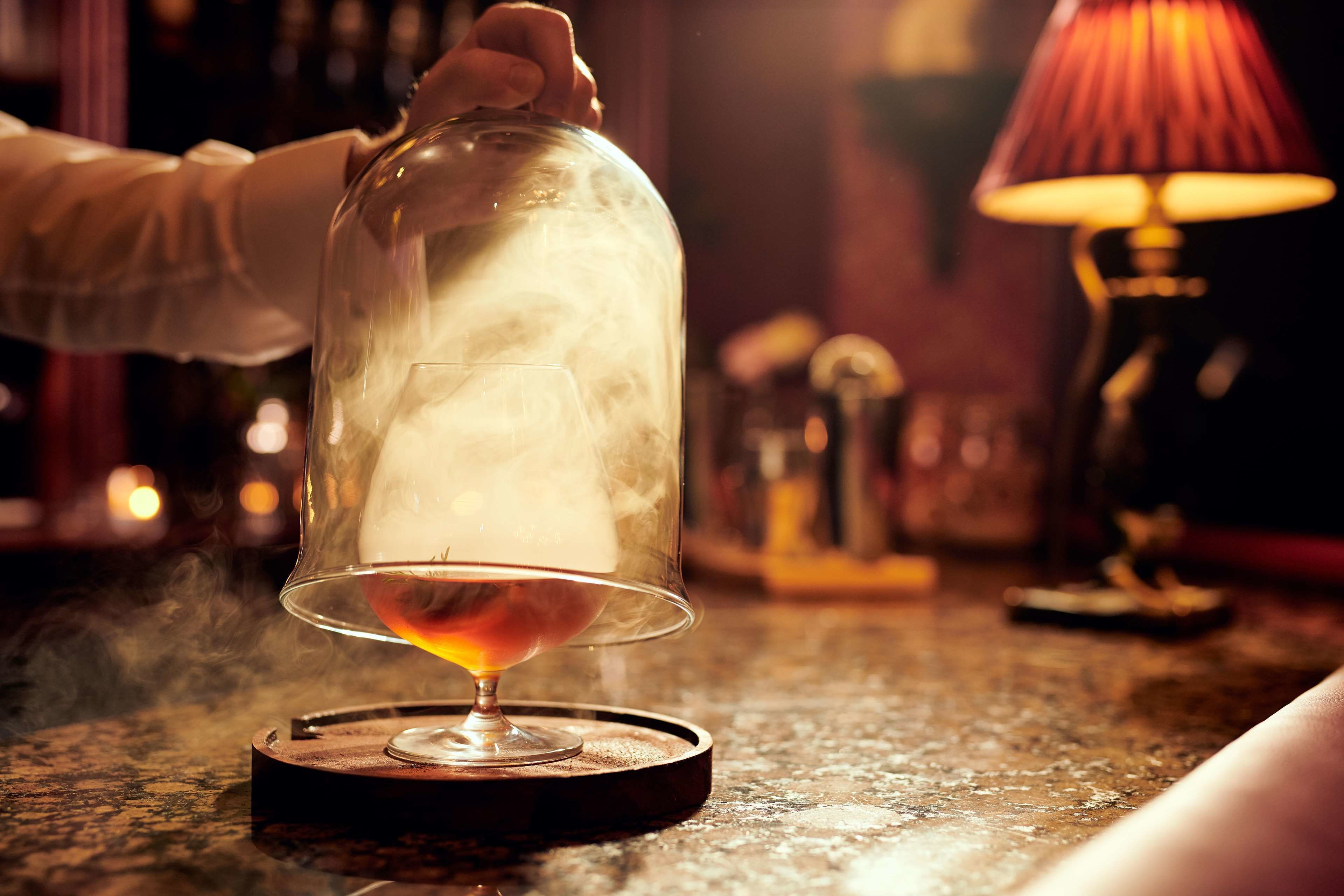 A hand lifting a glass dome over a candle, with a warm lamp in the background.