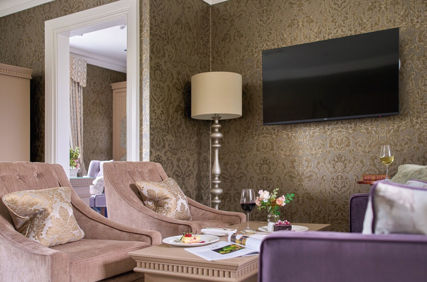 Habitación de hotel con un conjunto de sofás, una mesa con una copa de vino y un pequeño ramo de flores, y un televisor montado en la pared.