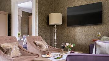 Habitación de hotel con un conjunto de sofás, una mesa con una copa de vino y un pequeño ramo de flores, y un televisor montado en la pared.