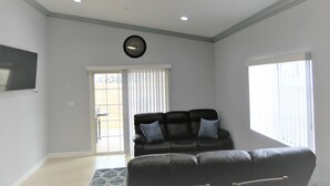 Een woonkamer met een zwarte leren bank, een klok aan de muur en een groot raam.