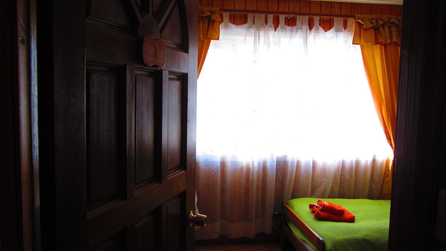 A room with a wooden door, a bed with a green bedspread, and orange curtains.