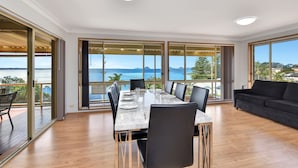 Un espace repas moderne avec une table dressée pour six personnes, de grandes fenêtres offrant une vue sur la mer et un balcon doté d’une terrasse en bois.