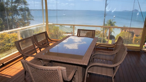 Un coin repas avec une table en verre et des chaises en osier, donnant sur un plan d’eau et une végétation luxuriante.