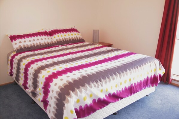 A bed with a patterned bedspread and pillows, a bedside table, and a red curtain. - Seaside Serenity Soldiers Point (Soldiers Point)