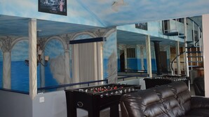 A room with a foosball table, a sofa, and a large mirror reflecting the room.