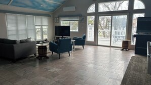 A spacious living room with a large TV, comfortable seating, and a view of the outdoors through large windows.