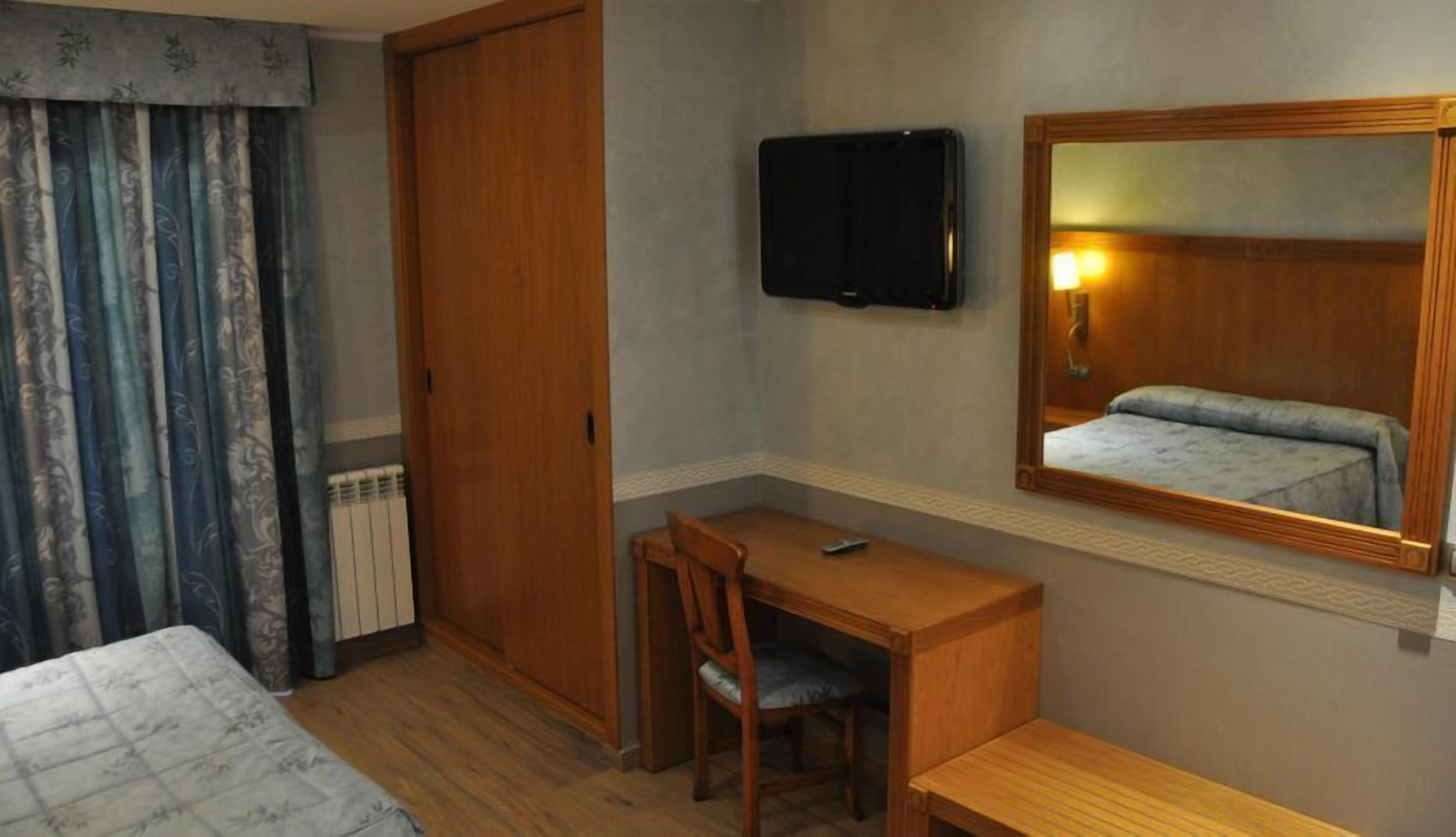 A hotel room with a wooden desk, a chair, a bed, a television, and a mirror.