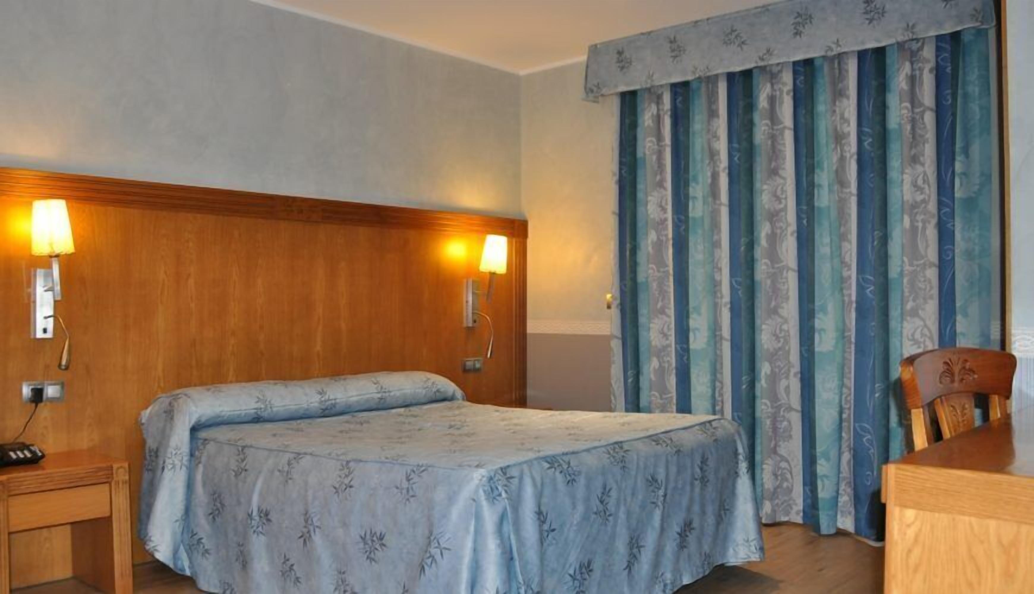 Habitación de hotel con cama doble, dos lámparas de noche, cabecera de madera, mesita de noche de madera, una silla de madera y una ventana con cortinas a rayas azules y blancas.
