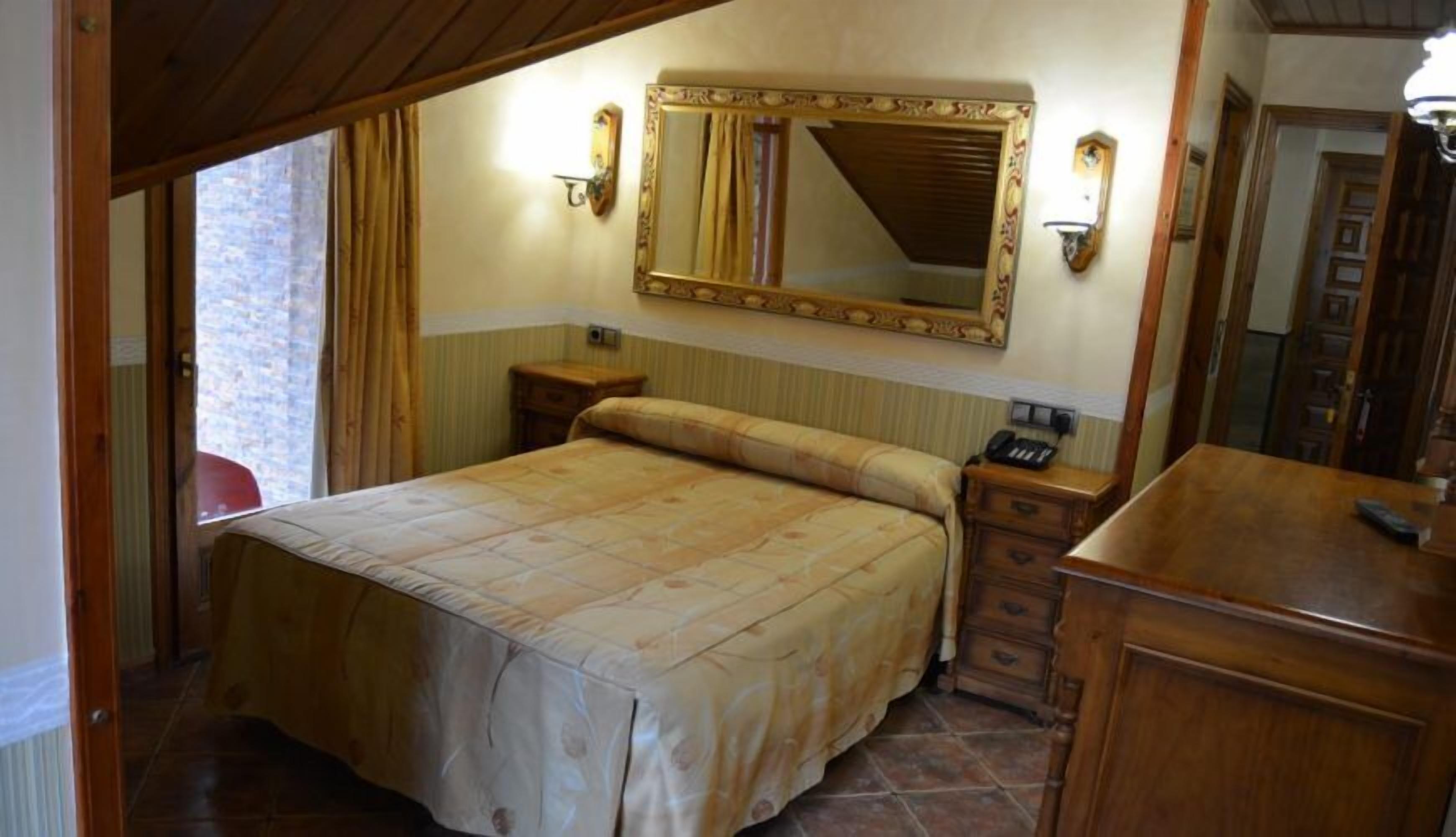 A bedroom with a large bed, a wooden dresser, a nightstand with a phone, and a mirror above the bed.