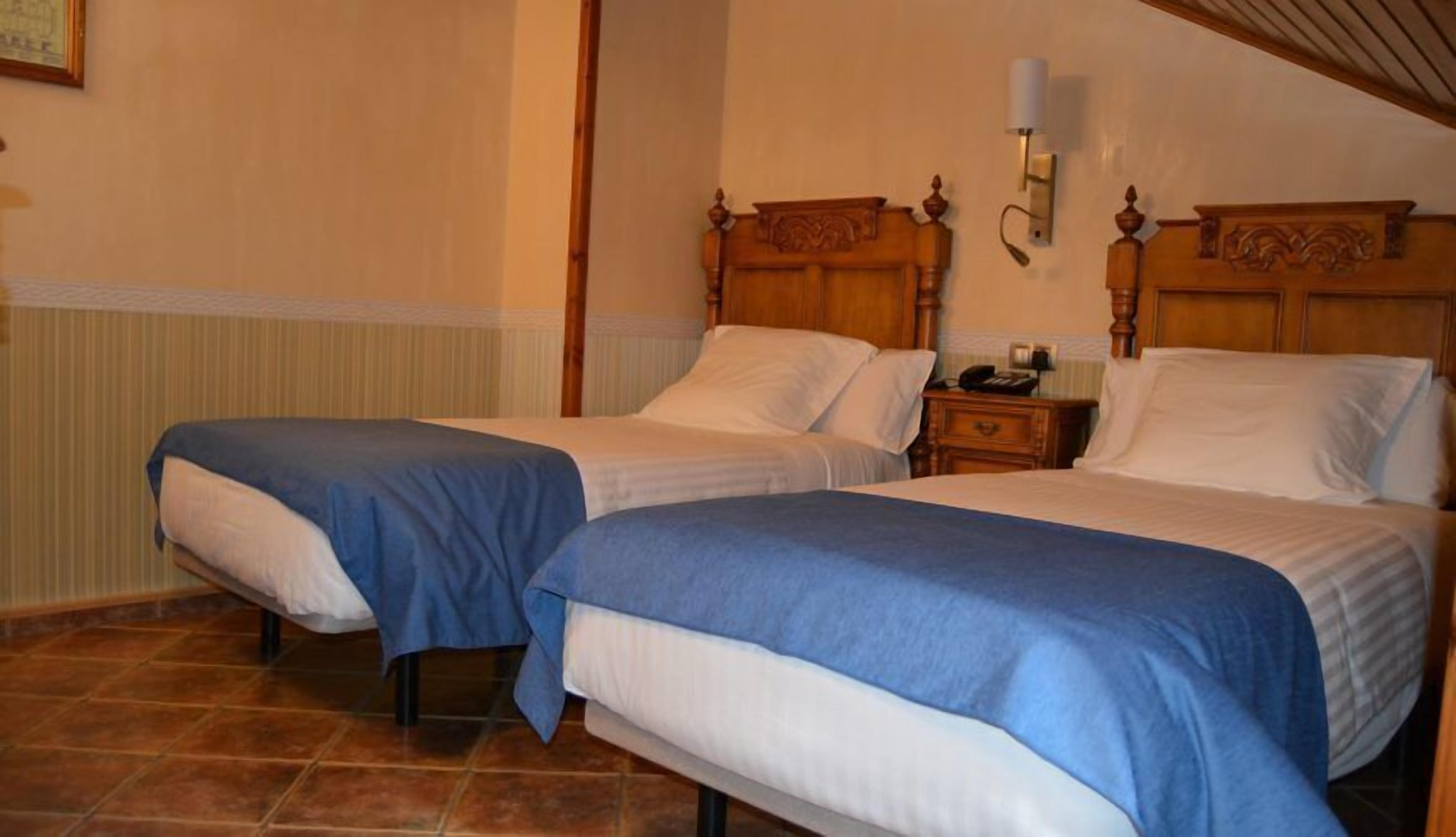 Una habitación de hotel con dos camas individuales, cabeceras de madera, una mesita de noche con lámpara y suelo de baldosas.