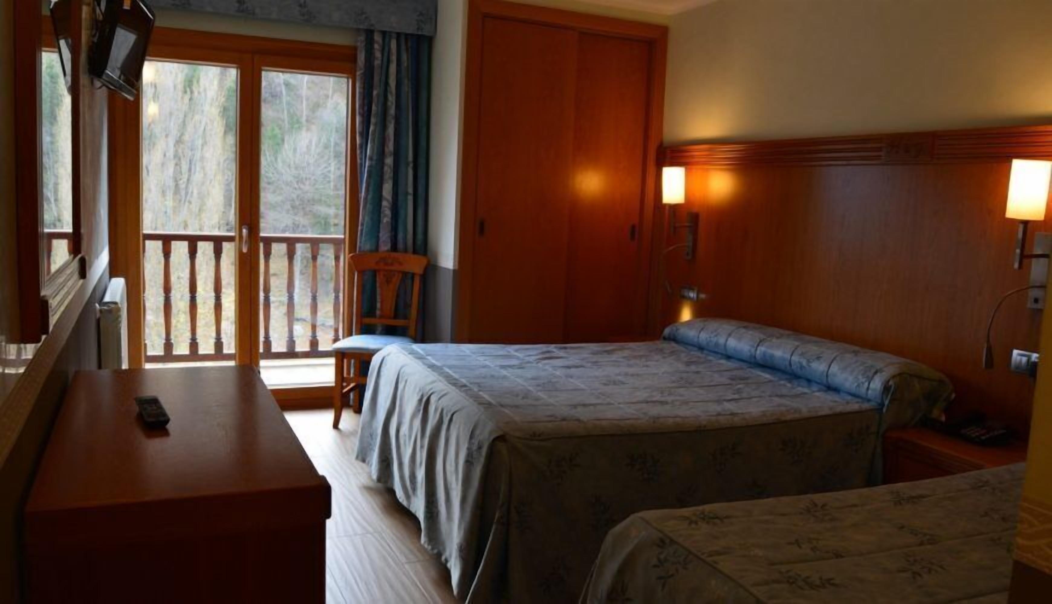 A hotel room with a bed, a desk, a chair, and a balcony with a view of trees.