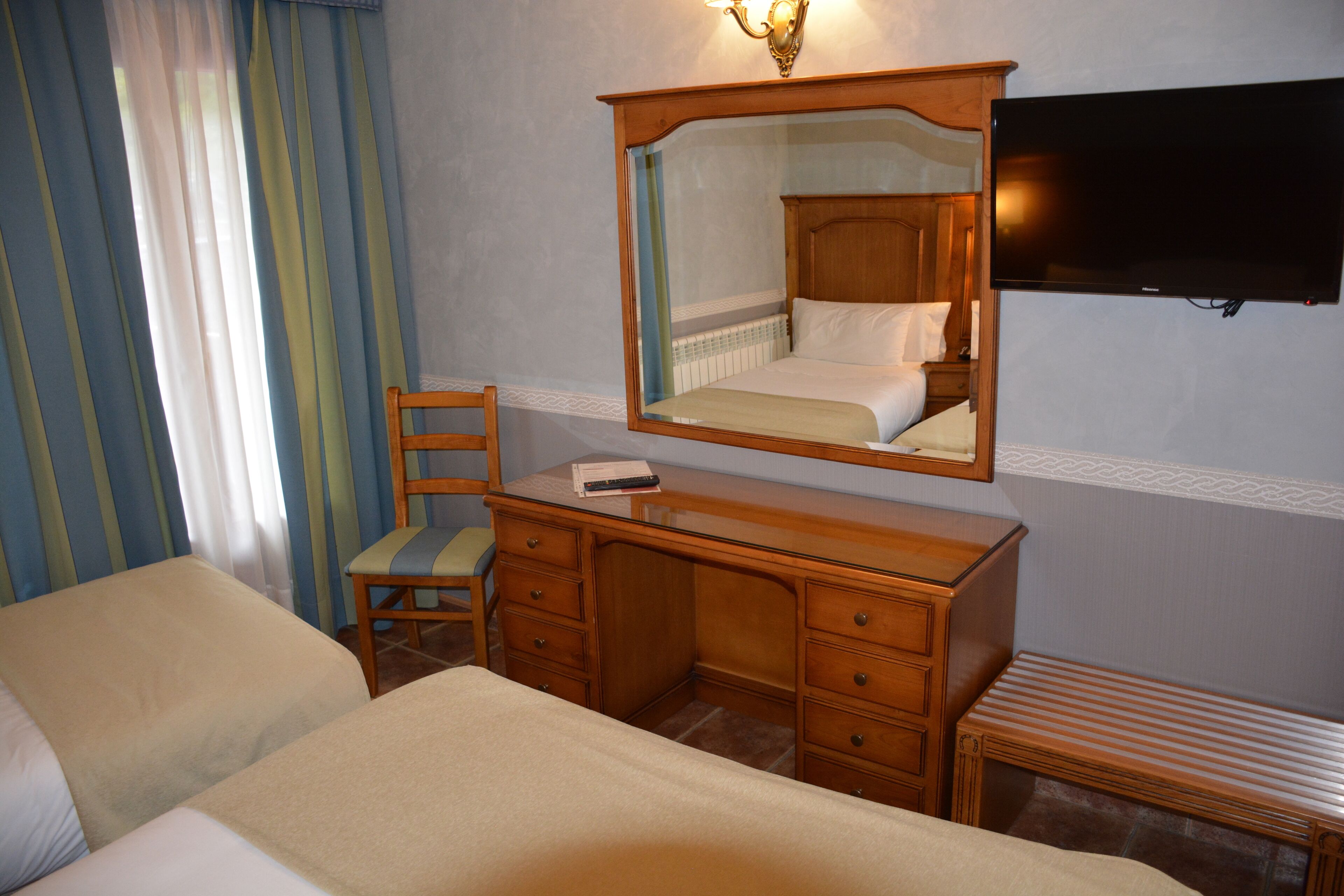 A hotel room with a bed, a wooden dresser, a chair, a television mounted on the wall, and a window with curtains.