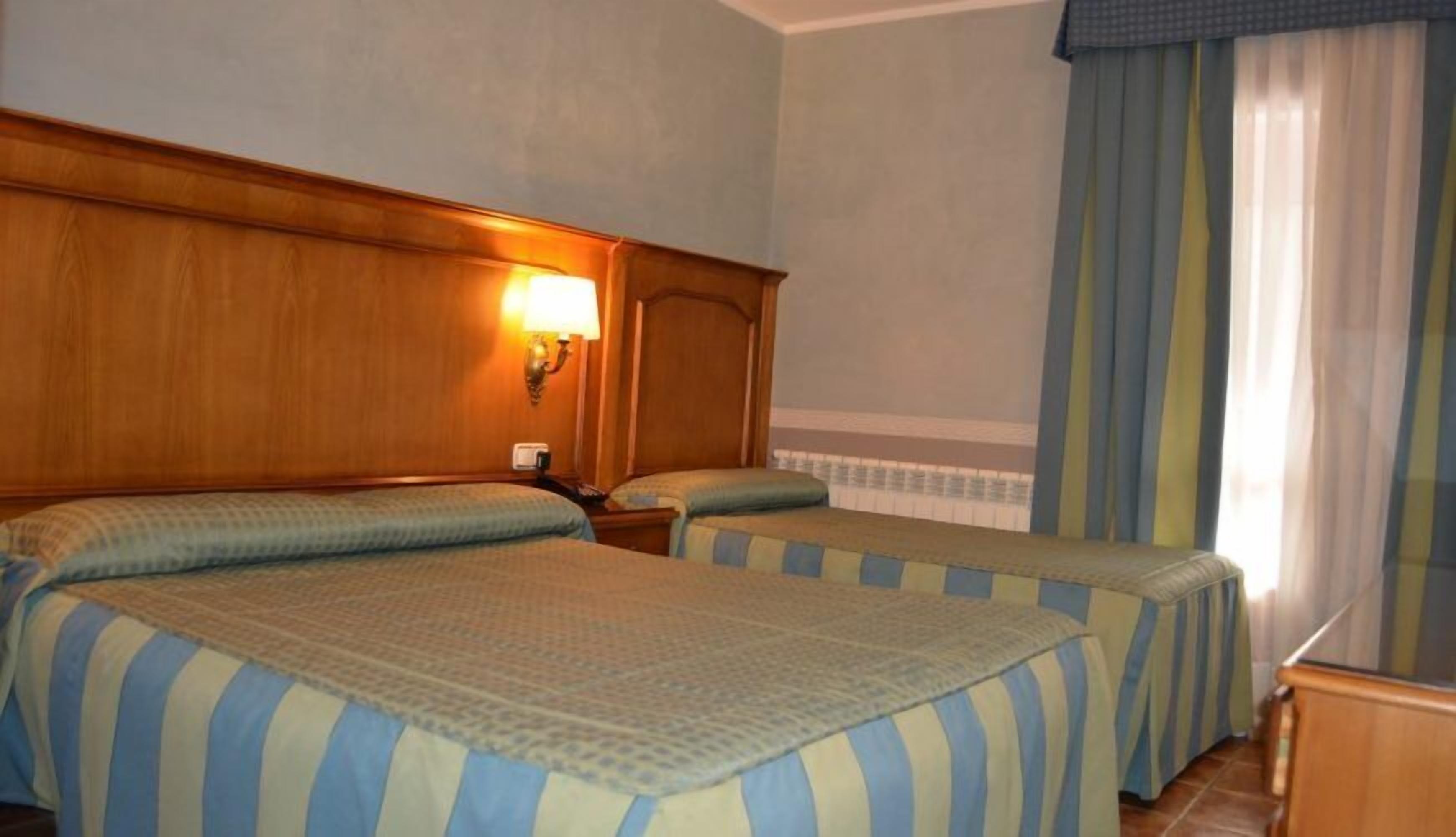 A hotel room with two single beds, a wooden headboard, a nightstand, a lamp, and a window with curtains.