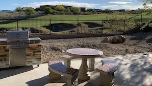 Eine Terrasse im Freien mit einem Grill aus Stein, einem runden Tisch mit Stühlen und Blick auf ein umzäuntes Grundstück mit Bäumen und ein Gebäude in der Ferne.