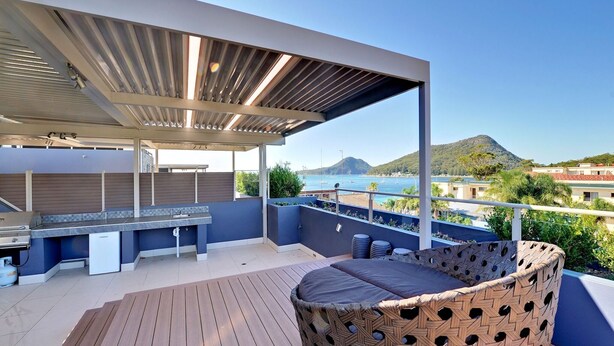 A covered outdoor area with a barbecue, seating, and a view of the sea and hills.