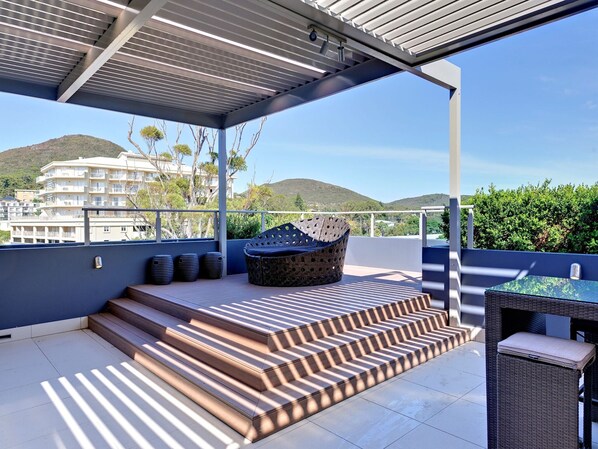 A covered outdoor area with steps leading to a seating area, a table, and a view of a hillside.