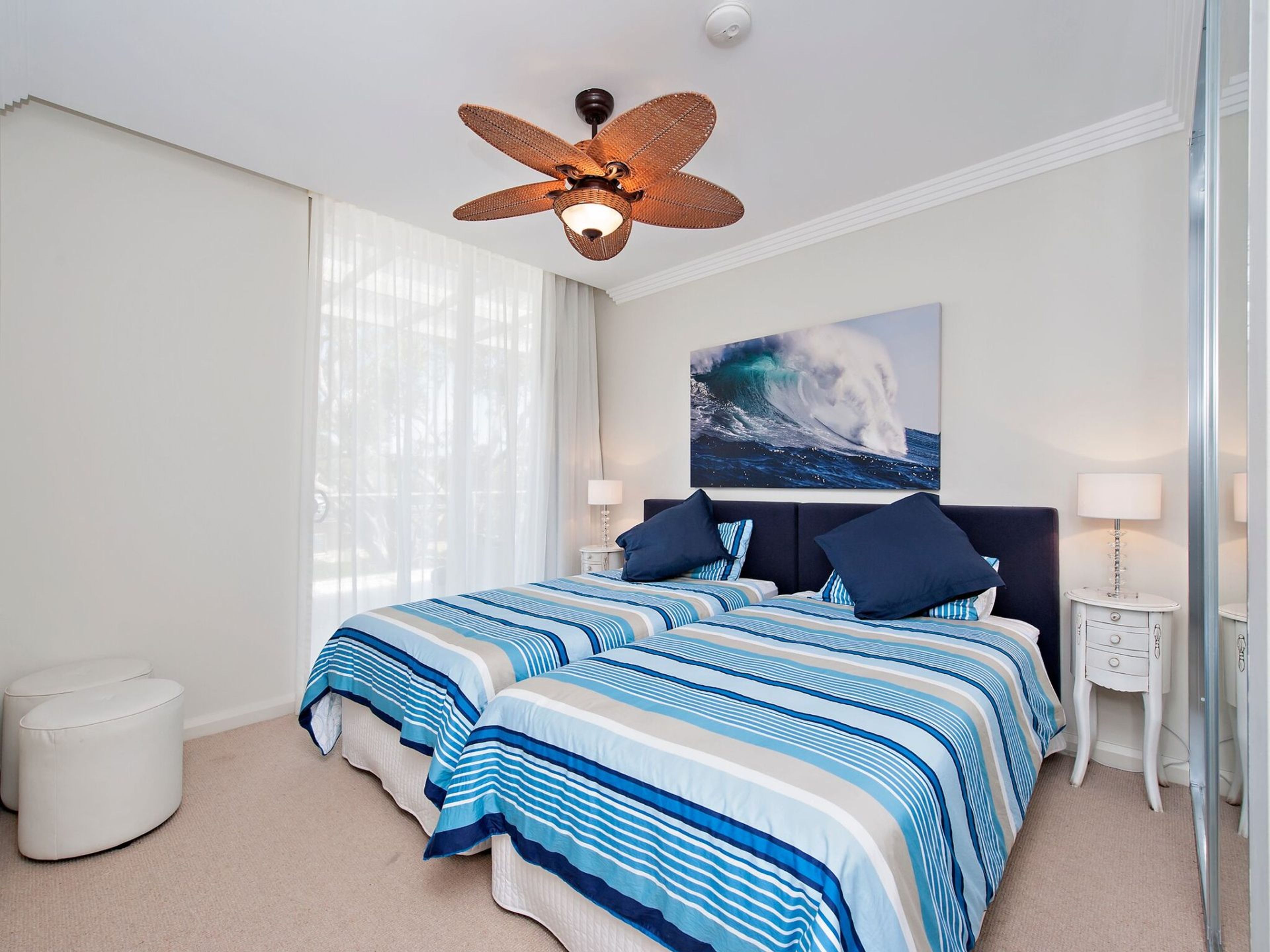 A bedroom with a large bed, a ceiling fan, a painting of a wave, and a nightstand with a lamp.
