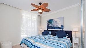A bedroom with a large bed, a ceiling fan, a painting of a wave, and a nightstand with a lamp. - Aura Apartments U12 Shoal Bay Beach Views (Shoal Bay)