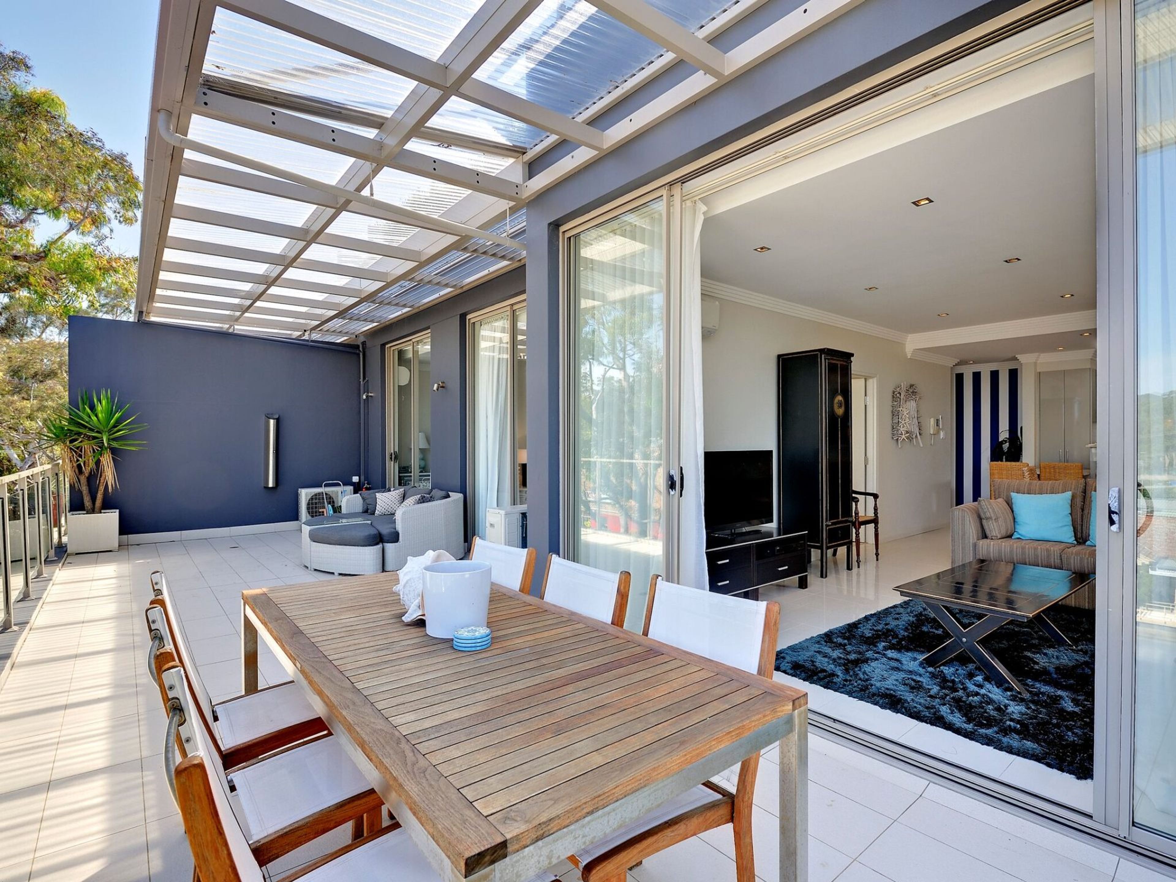 A modern living space with a wooden dining table, a sofa, and a television in a well-lit room with large windows.