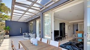 A modern living space with a wooden dining table, a sofa, and a television in a well-lit room with large windows. - Aura Apartments U12 Shoal Bay Beach Views (Shoal Bay)