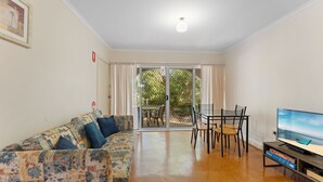 A living room with a sofa, a dining table with chairs, a TV, and a sliding glass door leading to an outdoor area. - Beach break garden unit! (Shoal Bay)