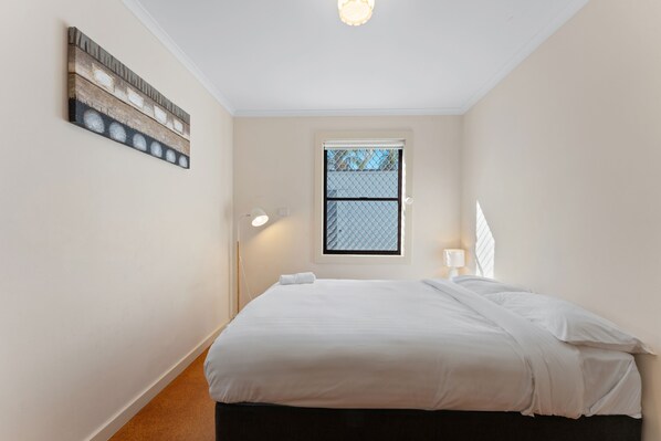 A bedroom with a bed, a window, a wall-mounted light, and a painting.